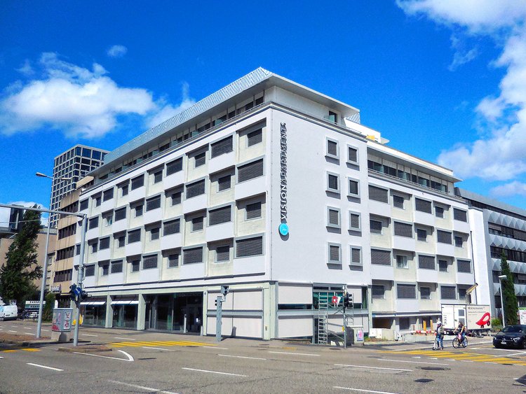 Photo: a modern, seven storey building with a light-grey façade and the VISIONAPARTMENTS sign, directly in front of the building a street crossing
