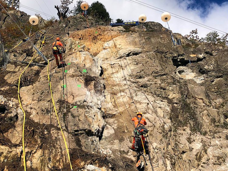 Foto: zwei Fels-Team Arbeiter am hängenden Seil in einer Felswand; am oberen Hang installierte Baustellenbeleuchtung für Nachtarbeiten