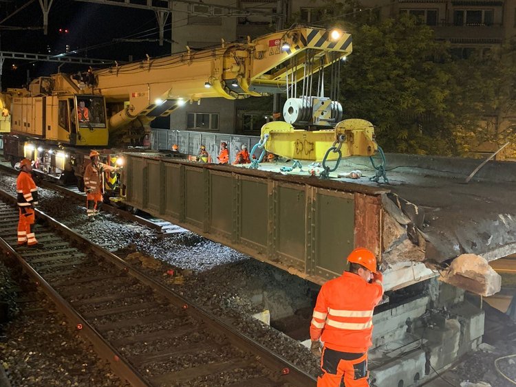 Foto: Das alte Brückenelement wird mit einem Kran abgehoben. Die Mitarbeiter in oranger Uniform stehen auf den Gleisen und betrachten das Vorgehen. 