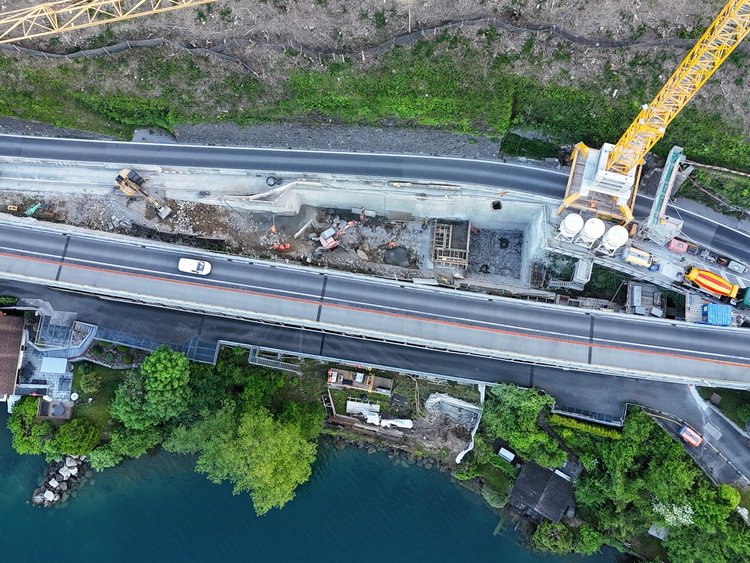 Foto: Luftaufnahme von der Baustelle mit grossem Kran. Die Achsenstrasse ist von Autos befahren und befindet sich zwischen See und der steilen Felswand. 