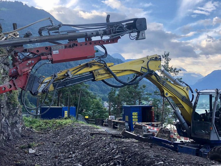 Photo: anchor drilling unit in operation in steep terrain; in the background Lake Uri, mountains, cloudy sky